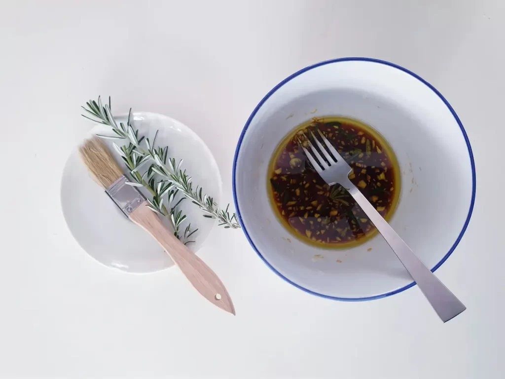 A bowl of dark brown marinade with herbs and garlic cloves, with a fork resting in it. Next to the bowl is a small white plate with a basting brush and a sprig of rosemary. All items are on a white surface.