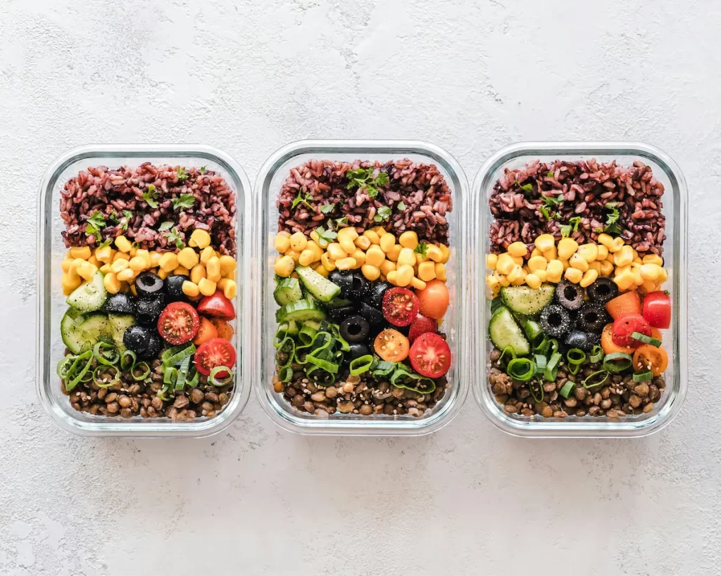 Three rectangular glass meal prep containers, each filled with layers of food. From top to bottom, each container has a layer of dark rice, yellow corn, sliced black olives, sliced cucumbers, and cherry tomatoes, with a base layer of lentils. The containers are arranged horizontally on a light, textured surface.